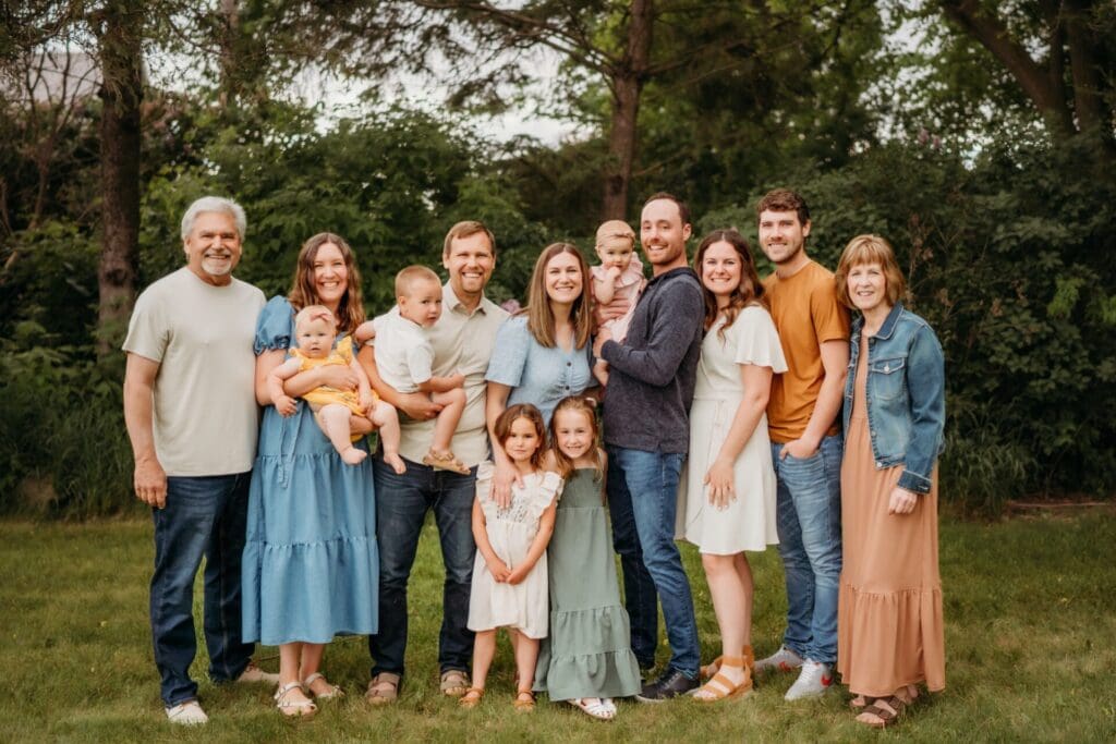 A large family poses together outdoors, smiling. Cindy Swenson captures six adults, three small children, two young girls, and a baby in casual summer clothes standing on grass with trees and bushes in the background.