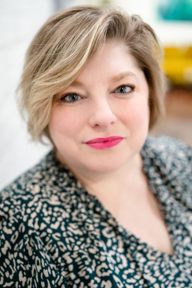 Angela Rantasha, a woman with short, wavy blond hair and pink lipstick, looks at the camera in a patterned black and white top. The softly blurred background reveals hints of white and yellow.
