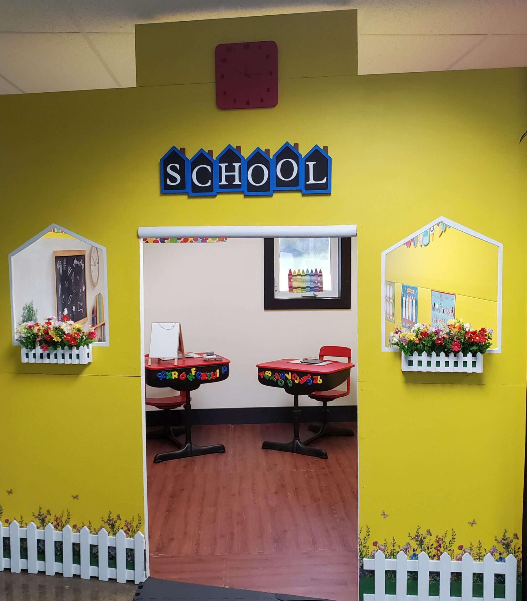 A yellow wall in Grammy's Town is styled like a school entrance, featuring a red clock, "SCHOOL" sign, white fences, flower boxes, and an inviting classroom with desks and colorful decor inside the indoor playground.