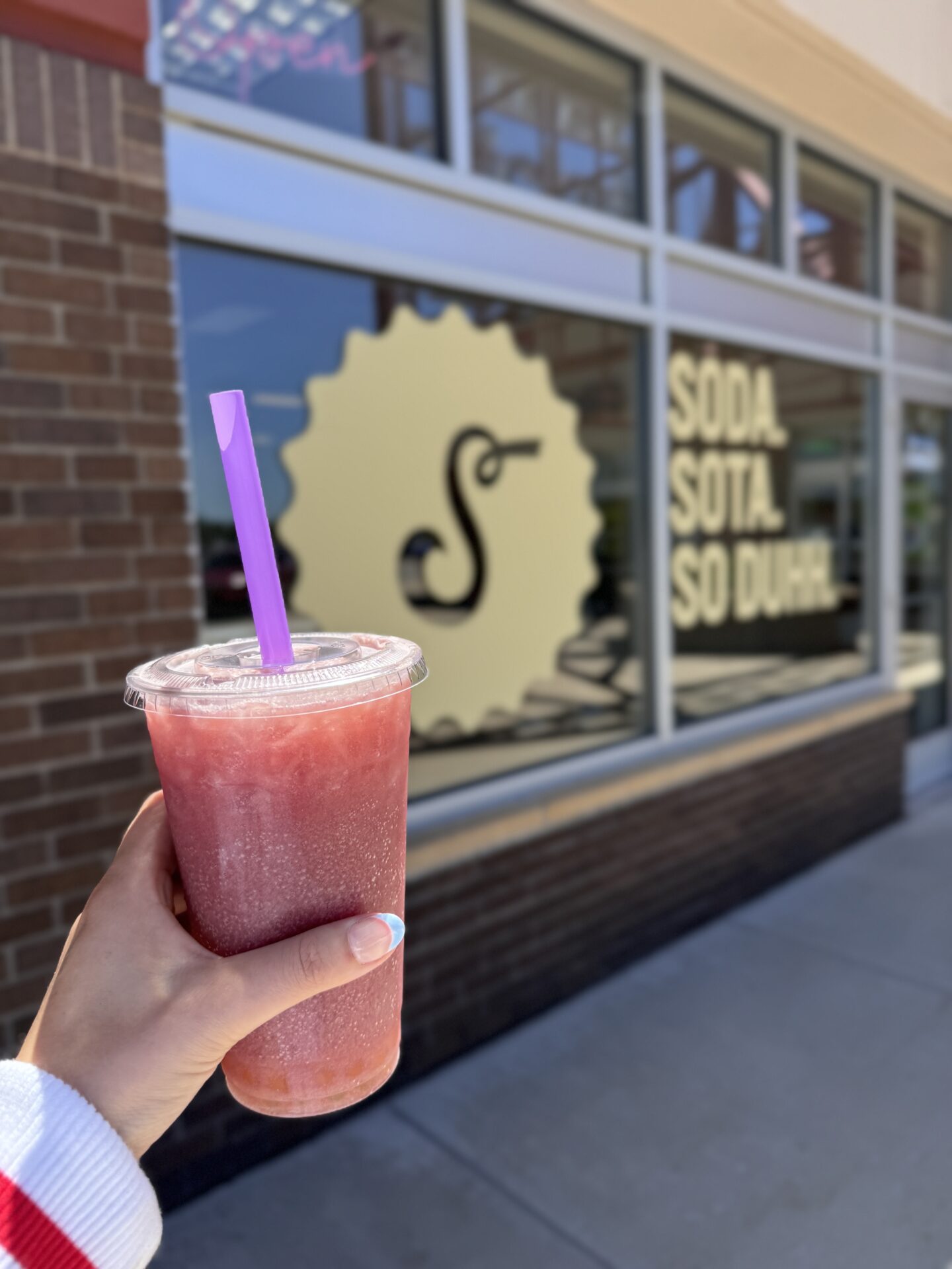 A hand with colorful nails holds a red slushy drink with a purple straw in front of Schmidty's Soda Shop window, displaying the text “SODA SOTA SO DUH!” and a stylized logo.