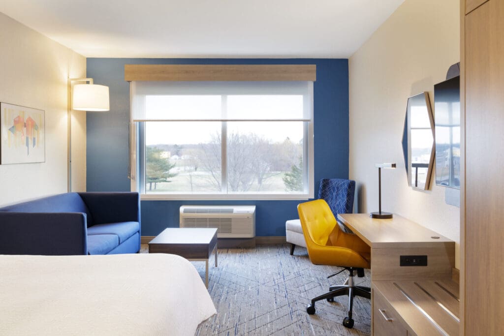 A modern Holiday Inn Express hotel room with a blue accent wall, a sofa, a work desk with a yellow chair, an armchair, and a large window overlooking trees and a grassy area.
