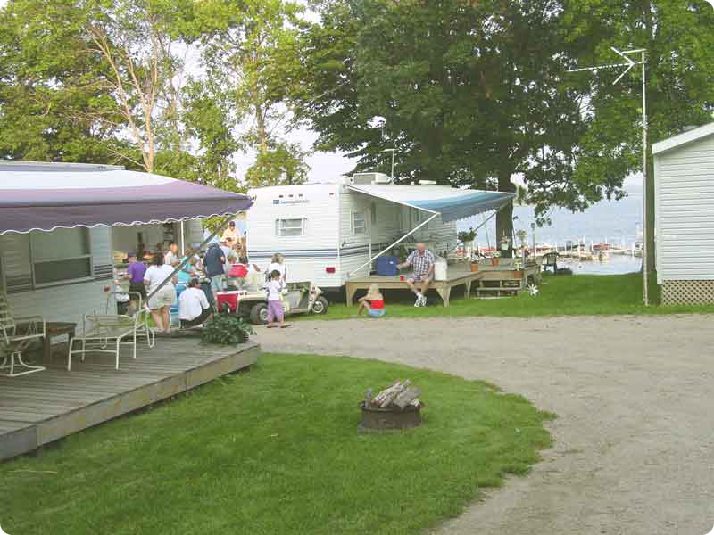People gather outside RVs at Big Foot Resort Campground, sitting at picnic tables under awnings. Trees, green grass, and a fire pit fill the foreground, while a lake or river shimmers in the background.
