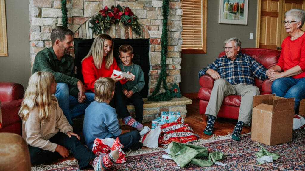 A family sits together in a cozy living room, exchanging and opening gifts in front of a decorated fireplace. As they enjoy the moment, their laughter suggests excitement to explore Alexandria MN after the holiday celebrations.