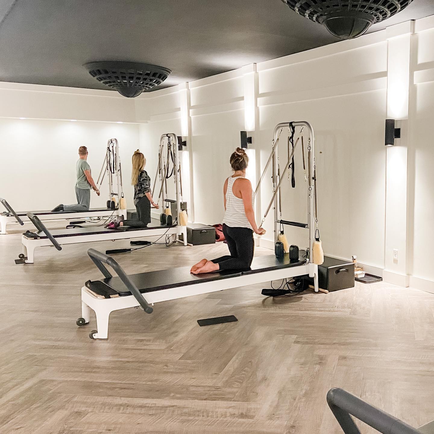Three people exercise on Pilates reformer machines in a bright, spacious studio with wooden floors, white walls, and modern lighting. At Cutting Edge Pilates, two women and one man each work at their own station for a balanced workout.