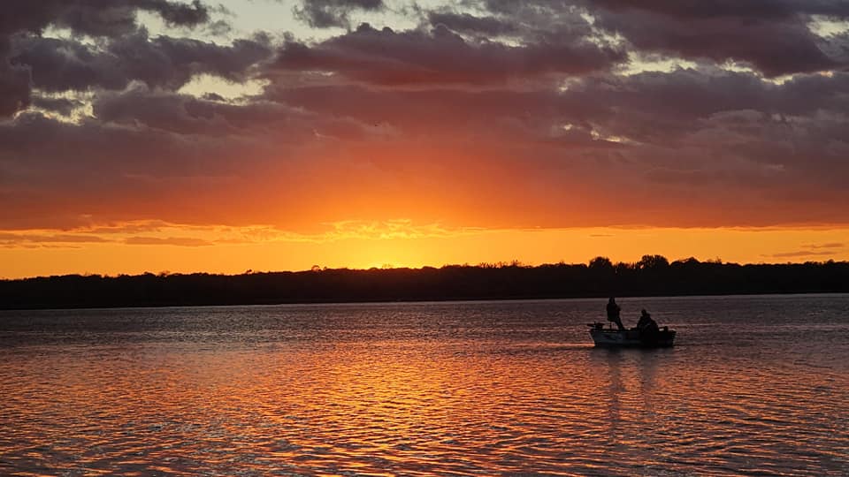 A small boat with two people floats on a calm lake at Eden Acres Resort during a vibrant orange and yellow sunset, with dark clouds overhead and the water reflecting the colorful sky.