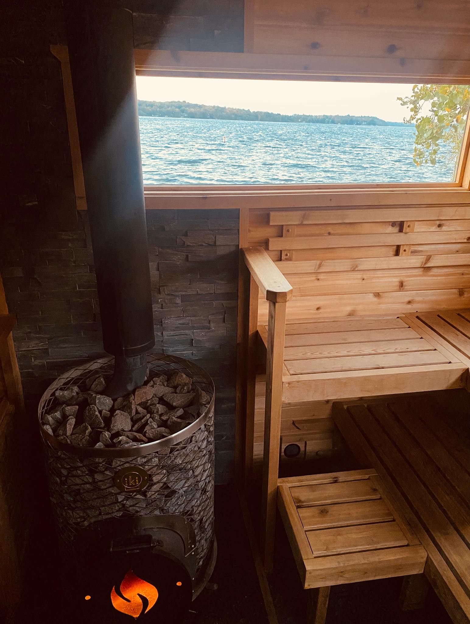 A cozy wood sauna with a stone stove and chimney is pictured indoors. A large window offers a scenic lake view, perfect for relaxing after a Cold Plunge. Wooden benches and walls create a warm, rustic atmosphere reminiscent of Northern Lights Sauna retreats.