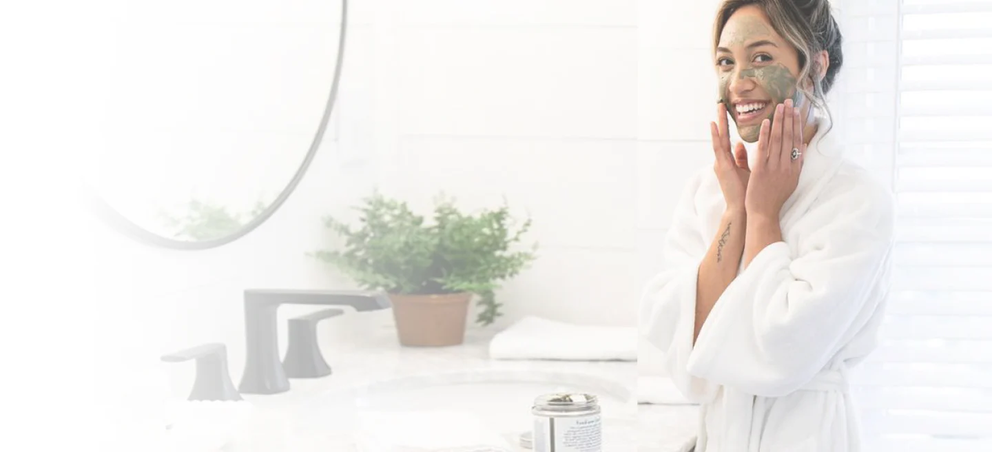 A smiling woman in a white bathrobe applies a green face mask during her DIY Facial Care routine by the sink, with a potted plant and towels in the bright bathroom—perfect inspiration for your next Facial Care Party.