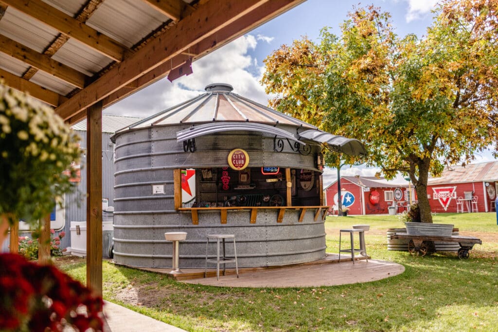 A round metal grain silo is converted into an outdoor bar with stools in front, set in a grassy area at a retro market, surrounded by trees and red barns under a partly cloudy sky.