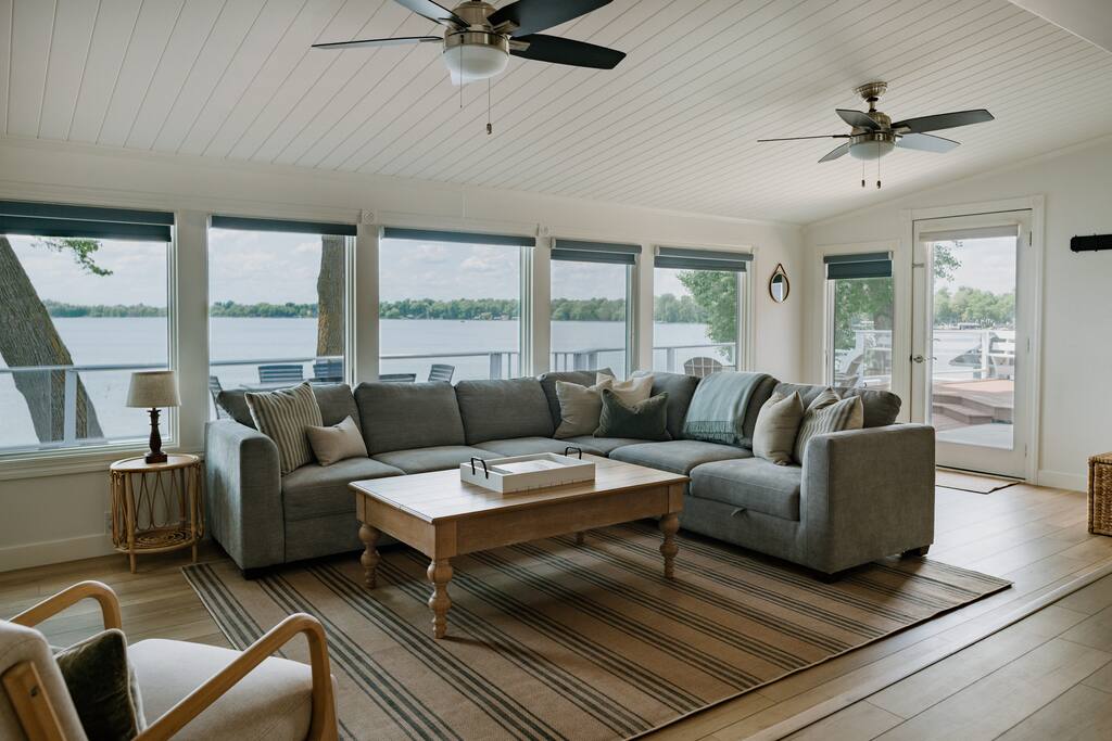 Bright living room at Alex Landing with large windows overlooking a lake, featuring a gray sectional sofa with cushions, a wooden coffee table on a striped rug, ceiling fans, a chair, and a door leading to the deck outside.