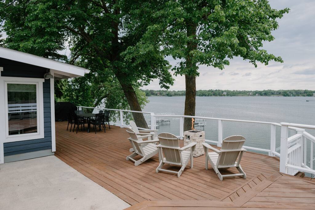 A spacious wooden deck at Alex Landing overlooks a calm lake, featuring white railings, Adirondack chairs around a fire pit, and an outdoor dining table under leafy trees beside a dark blue house.