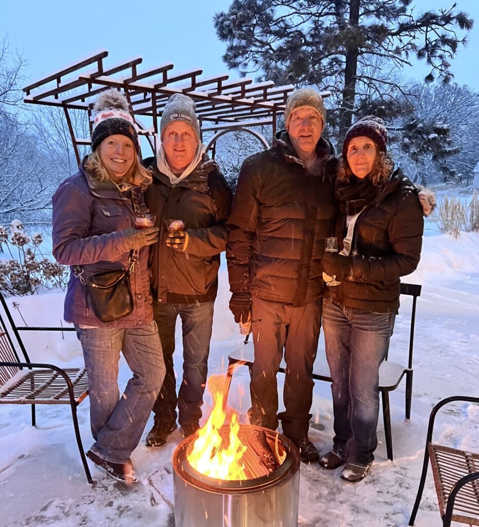 Four adults in winter clothing stand together outside around a fire pit in a snowy backyard, holding drinks and smiling at the camera—capturing the cozy spirit often found at Alexandria Winter Festivals. Snow blankets the ground, trees, and outdoor furniture nearby.