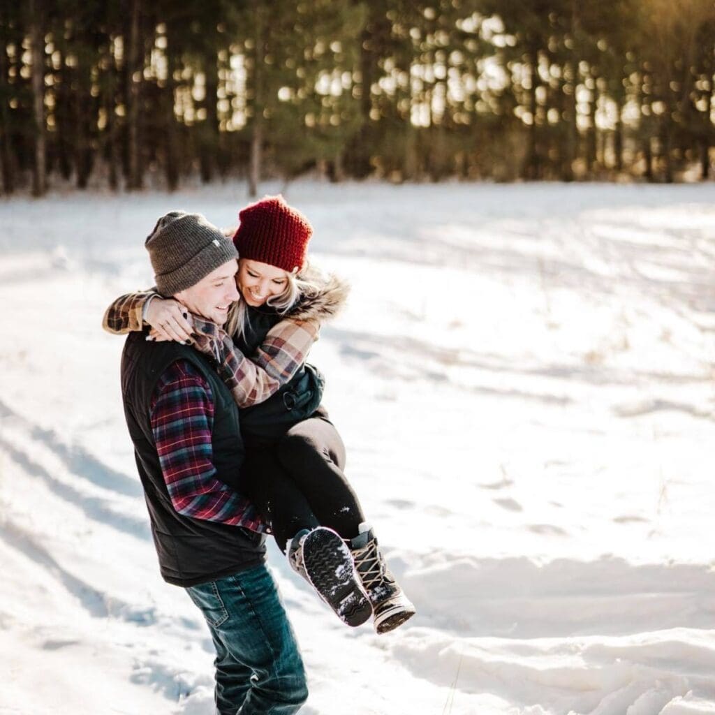 A man in winter clothes lifts a smiling woman wearing a red knit hat and boots in a snowy Alexandria MN field with pine trees behind them. Sunlight shines through, creating a warm, cheerful Valentine’s Day atmosphere.