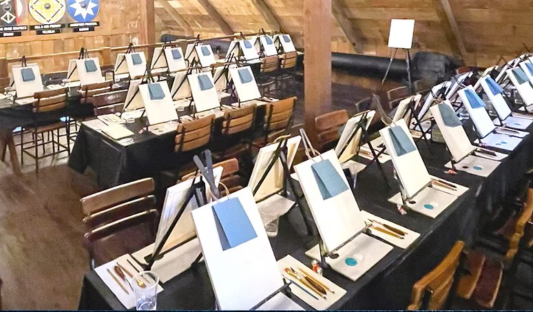 A room in the Palette District with rows of tables set up for a painting class, each featuring a canvas on an easel, paintbrushes, palettes, and cups. Wooden walls and a sloped ceiling complete the creative setting.