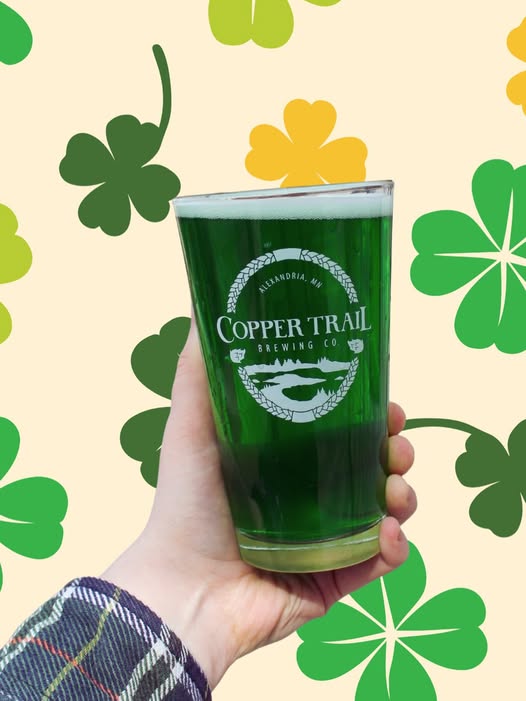 A hand holds a pint of green beer in a Copper Trail Brewing Co. glass, with green and yellow shamrocks in the background—perfect for celebrating St. Pats Weekend.