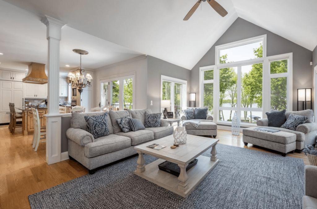 Spacious living room with gray sofas, a light wood coffee table, and large windows revealing Lake Le Homme Dieu—a true lakefront escape—open to a bright kitchen with white cabinets, wood range hood, and light hardwood floors throughout.