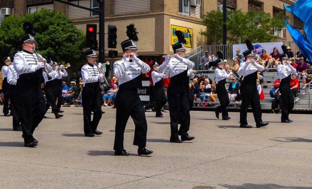 A marching band in black and white uniforms performs in a parade at the Annual Band Festival, playing instruments as people watch from bleachers and sidewalks in the background.