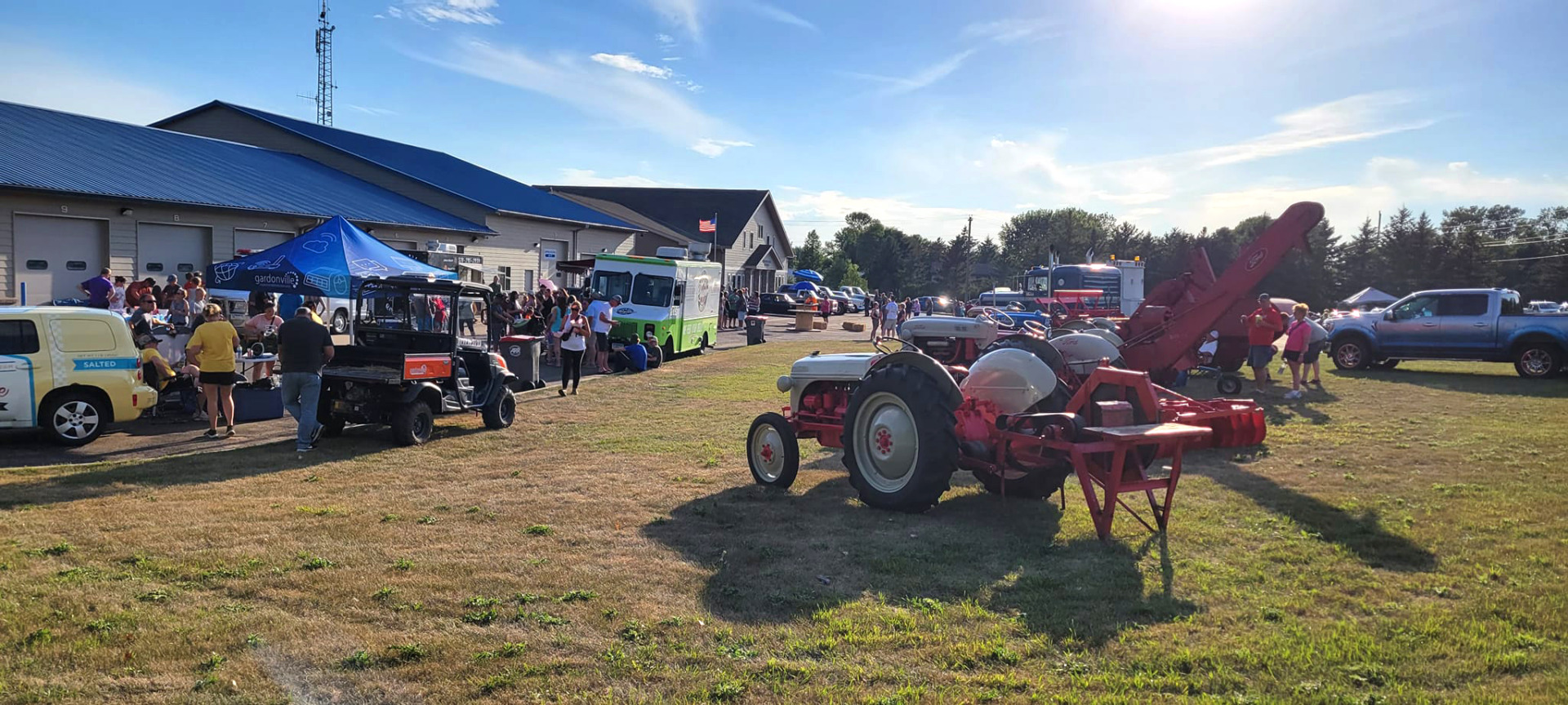 People gather outdoors at the Brandon Car Show, a community event featuring classic vehicles like a vintage tractor in the foreground, with food trucks, tents, and a building in the background on a sunny day.