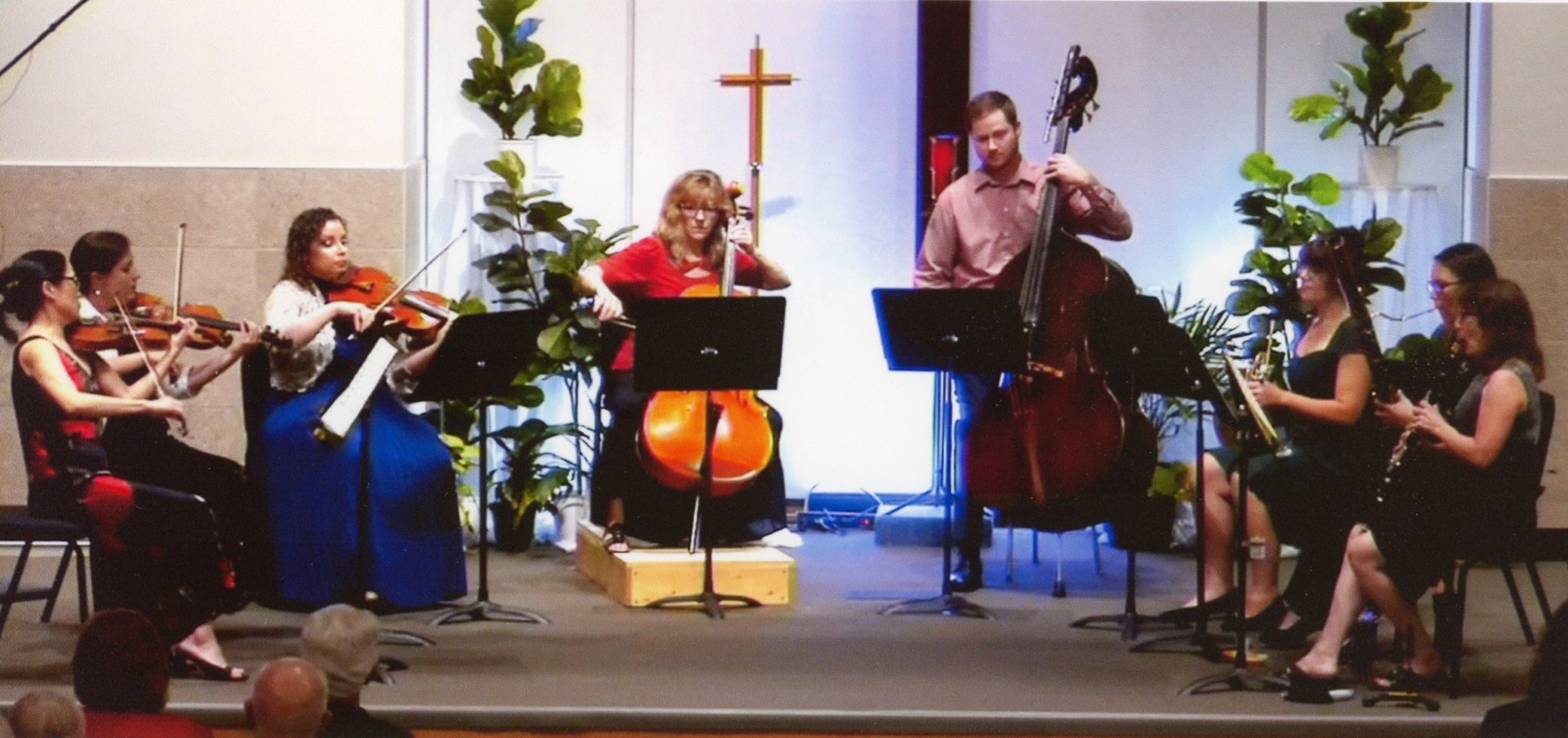 Seven musicians perform on stage in a church during the Alexandria Festival, playing string and woodwind instruments, with sheet music stands in front of them and green plants in the background.