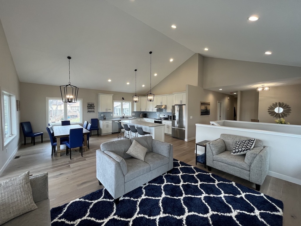 Open-concept living space at Le Homme Dieu Cove with modern kitchen, dining area, and living room. Features gray sofas, a navy geometric rug, blue dining chairs, large windows, pendant lights, and light wood floors.