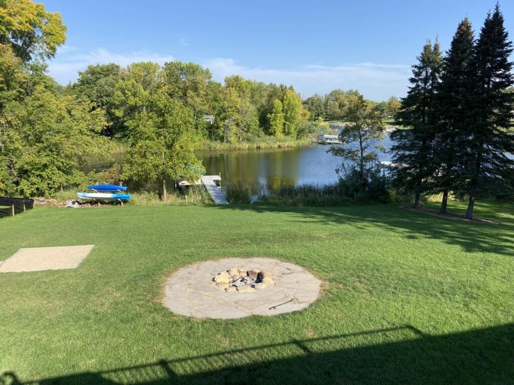 Lush green lawn with a stone fire pit in the foreground, a small lake and dock with boats in the middle ground, and trees surrounding Lake Le Homme Dieu under a clear blue sky near Crestwood Lodge.