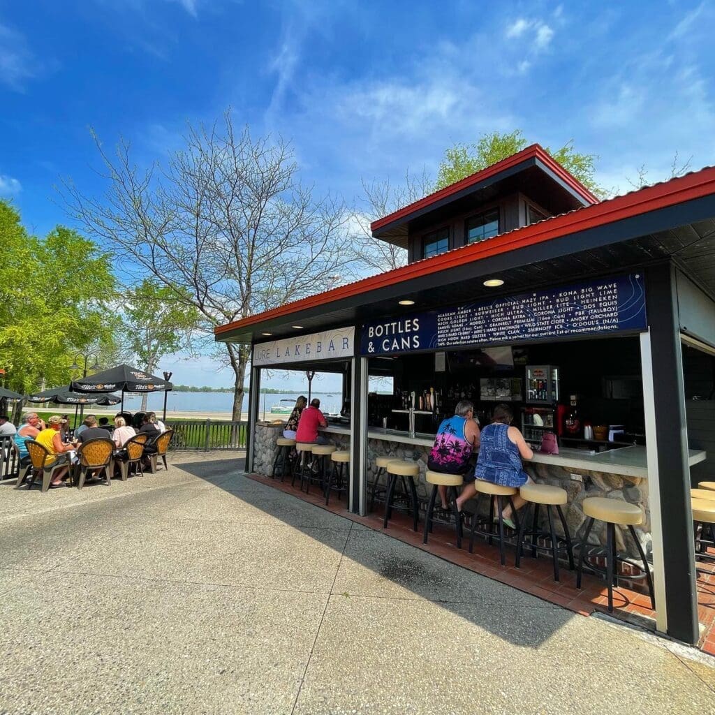 Outdoor bar, Lure Lakebar, with people sitting at the counter and nearby tables under a sunny blue sky. Trees with spring foliage surround the area, a lake is visible in the background, and live music adds to the lively atmosphere.