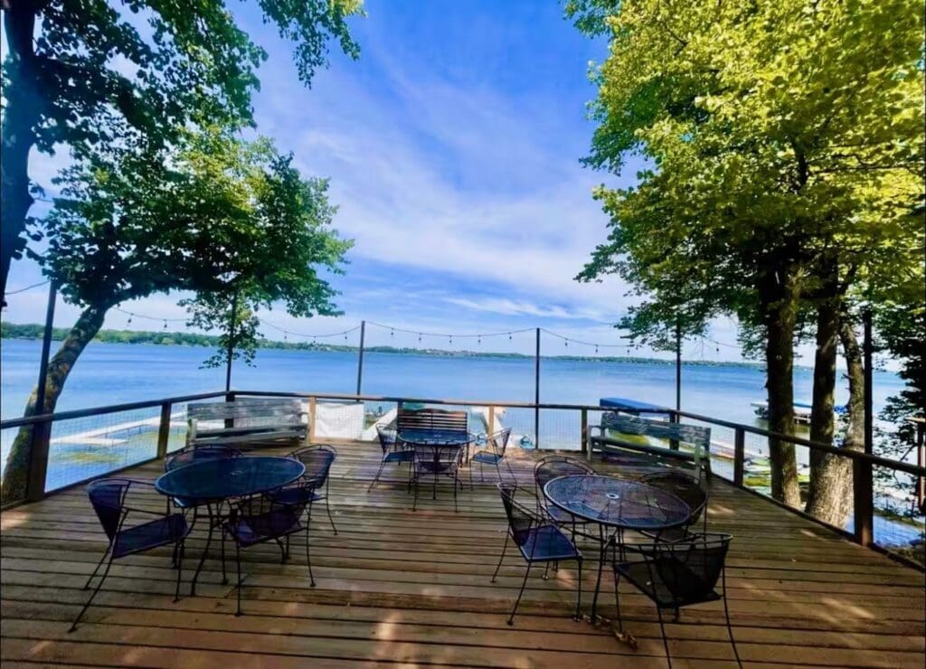 A wooden deck at The Nest with metal tables and chairs overlooks a calm lake at Darling Shores, framed by green trees and string lights under a blue sky. Benches line the railing, offering a scenic, peaceful view.