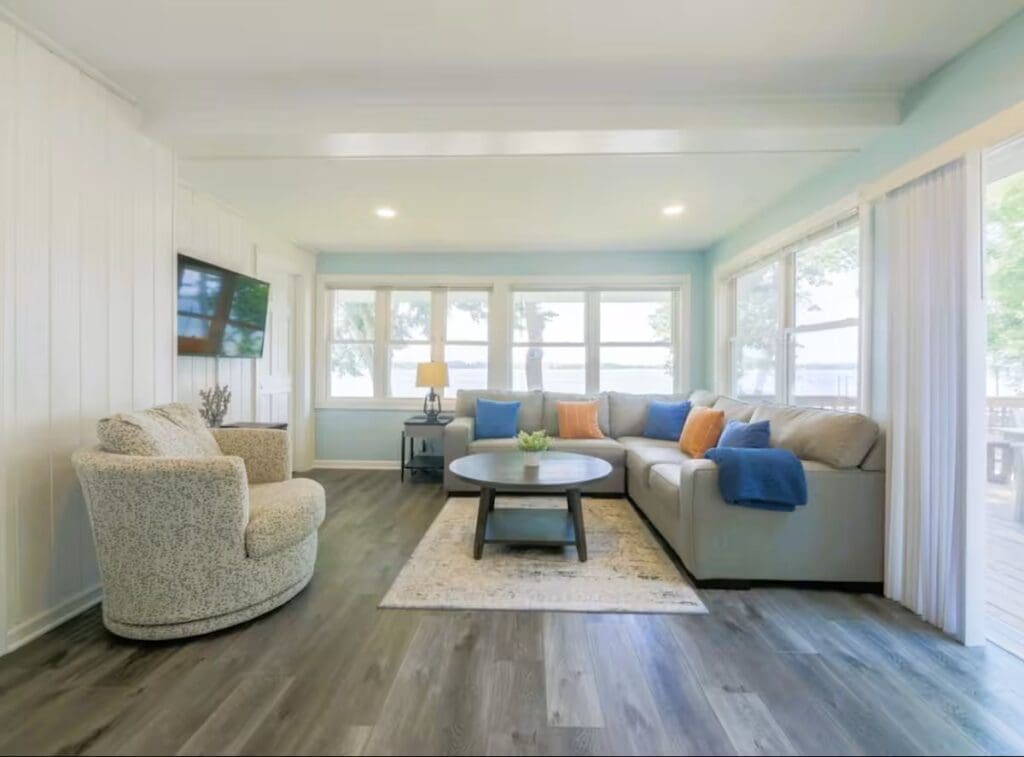 Bright living room at Darling Shores with large windows, an L-shaped sofa with blue and orange pillows, a round coffee table on a rug, swivel chair, wood floors, wall-mounted TV, and gorgeous Lookout views of trees and water.