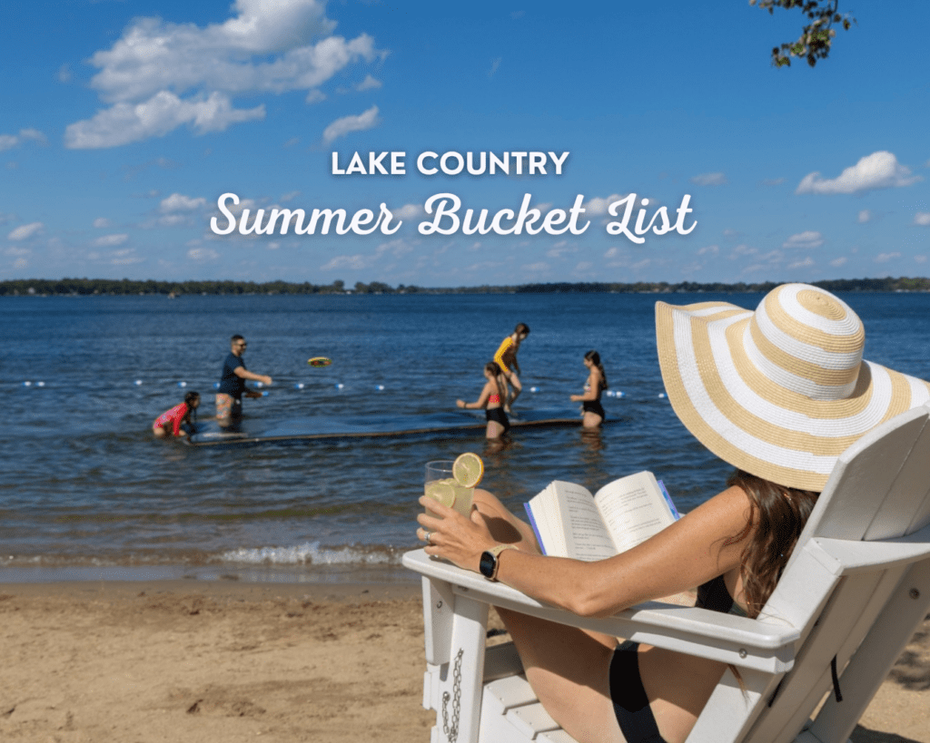 A woman in a sunhat relaxes on a beach chair with a book and a drink as people play in the lake behind her. Text reads: "Lake Country Summer Bucket List—Savor the Season." Blue sky with clouds overhead.