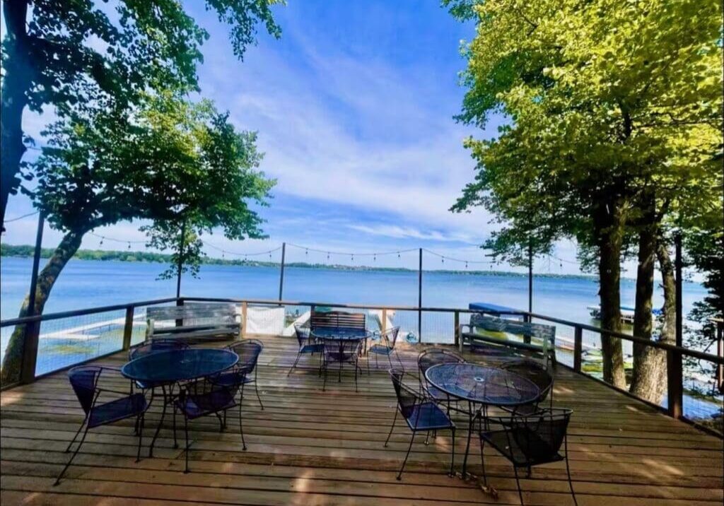 A wooden deck at The Nest with metal tables and chairs overlooks a calm lake at Darling Shores, framed by green trees and string lights under a blue sky. Benches line the railing, offering a scenic, peaceful view.