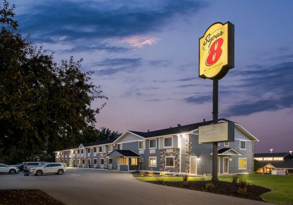 A Super 8 motel exterior at dusk features lit Super 8 signage, parked cars in front, and a tree framing the left side. The partly cloudy sky glows with subtle evening light.