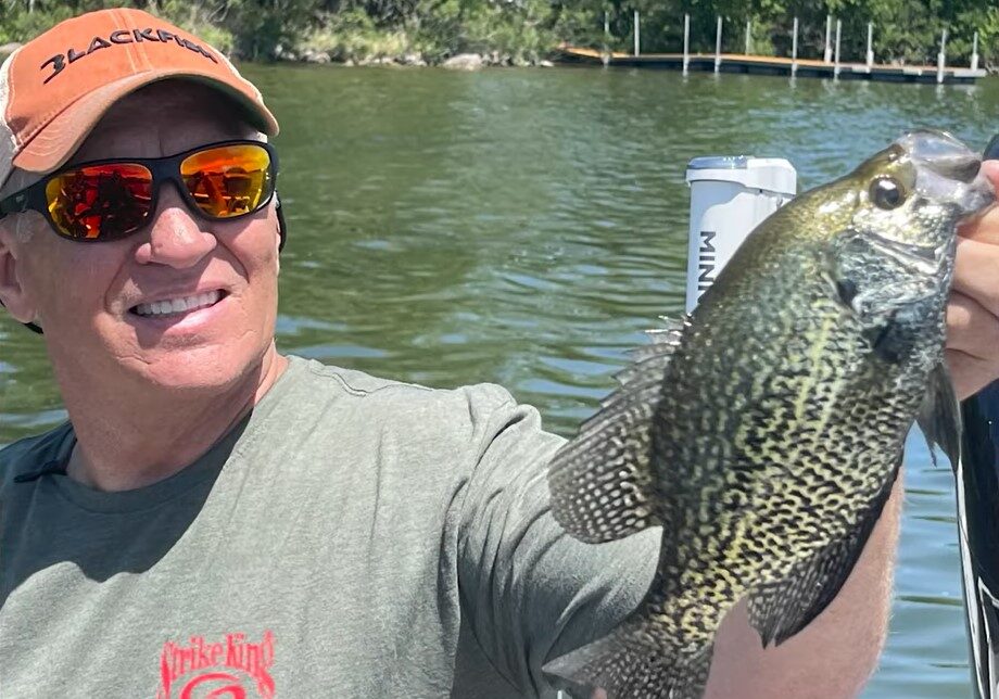 A smiling man in sunglasses and a cap holds up a large crappie fish on a boat, showcasing his success fishing the Midwest, with a lake and trees in the background.