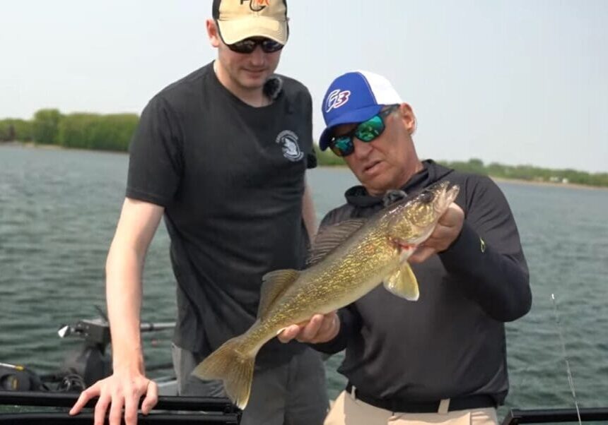 Two men on a boat, one in a black T-shirt and beige cap, the other in sunglasses and a blue cap, proudly hold an Alexandria walleyes catch with greenish-yellow scales over the water. Trees and shoreline can be seen in the background.