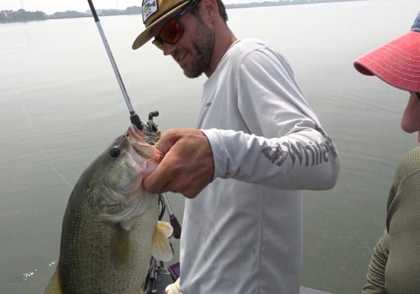 A man in sunglasses and a hat holds a large fish with one hand while standing on a boat, showcasing the thrill of fishing the Midwest. Another person in a red cap stands nearby, with calm water and a hazy shoreline in the background.