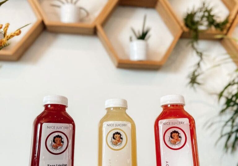 Three bottled drinks from Nice Juicery—labeled "Teaz Louise," "Mingle Gini," and "Pink Lady"—stand on a counter. Behind them, honeycomb-shaped wall shelves display plants and decor against a white wall.