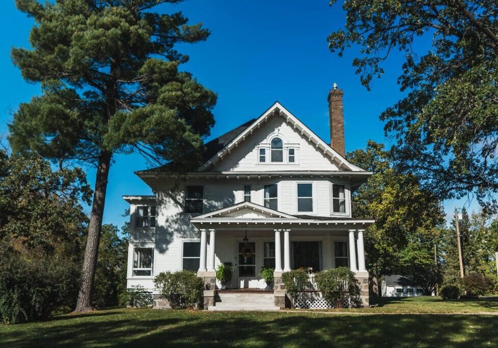 A large, white, two-story house—The Starling House—with a front porch and gabled roof sits on a grassy lawn, surrounded by tall trees under a clear blue sky, offering one of the most charming places to stay in Alexandria MN.