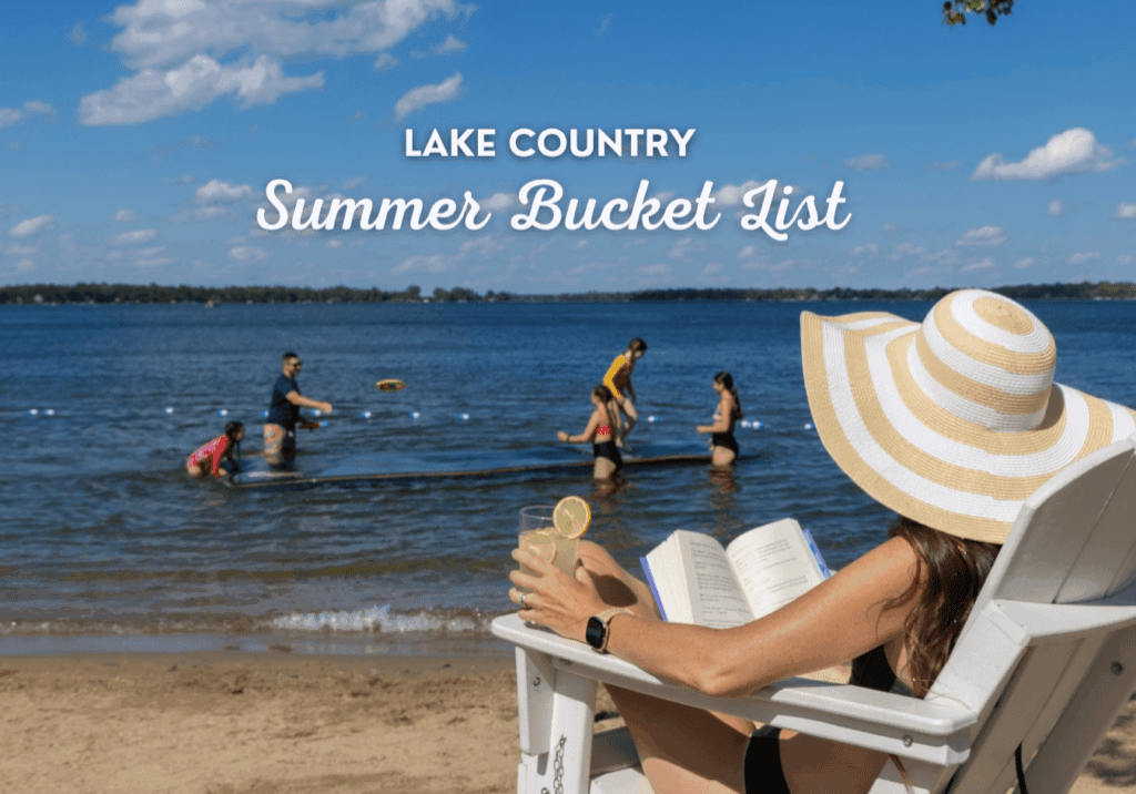 A woman in a sunhat relaxes on a beach chair with a book and a drink as people play in the lake behind her. Text reads: "Lake Country Summer Bucket List—Savor the Season." Blue sky with clouds overhead.