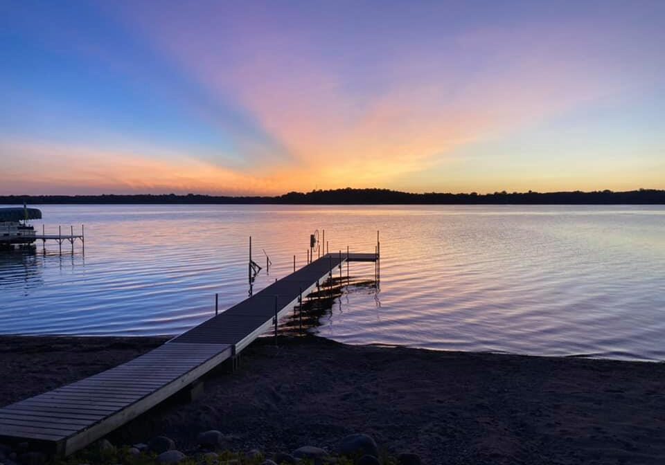 A wooden dock extends over calm lake water at sunset in Sandy Beach Haven, with vibrant orange, yellow, and purple hues lighting up the sky. Trees form a dark silhouette along the distant shoreline.