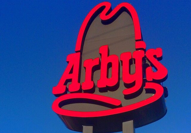 The image shows a brightly lit Arby's restaurant sign with bold red lettering and the iconic cowboy hat outline against a clear blue sky.