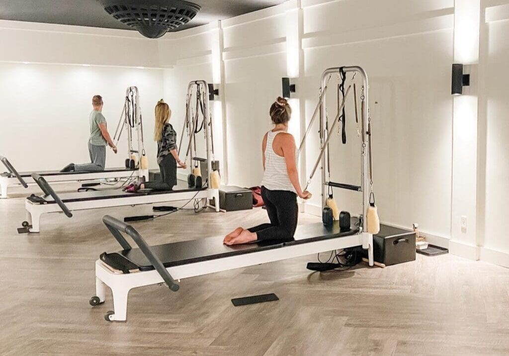 Three people exercise on Pilates reformer machines in a bright, spacious studio with wooden floors, white walls, and modern lighting. At Cutting Edge Pilates, two women and one man each work at their own station for a balanced workout.