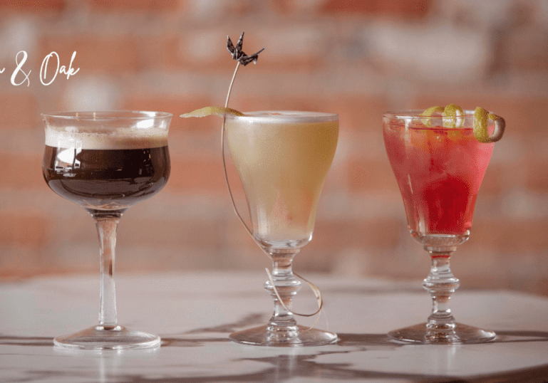 Three elegant cocktails from Tannin & Oak sit on a marble table: a dark cocktail with foam, a pale yellow drink garnished with a twist and flower, and a pink drink with lime slices. "Tannin & Oak" is written in the top left corner.