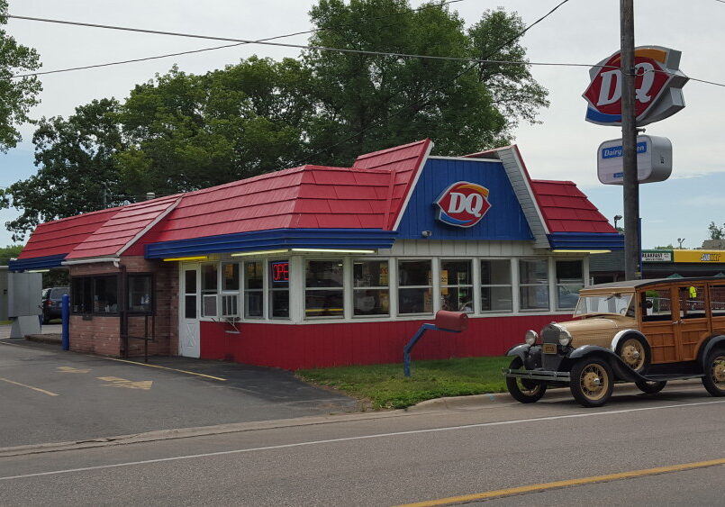 A Dairy Queen North restaurant with a red roof and blue trim sits near a road. An antique car is parked in front, and the DQ sign is visible above the building, with trees and other buildings in the background.