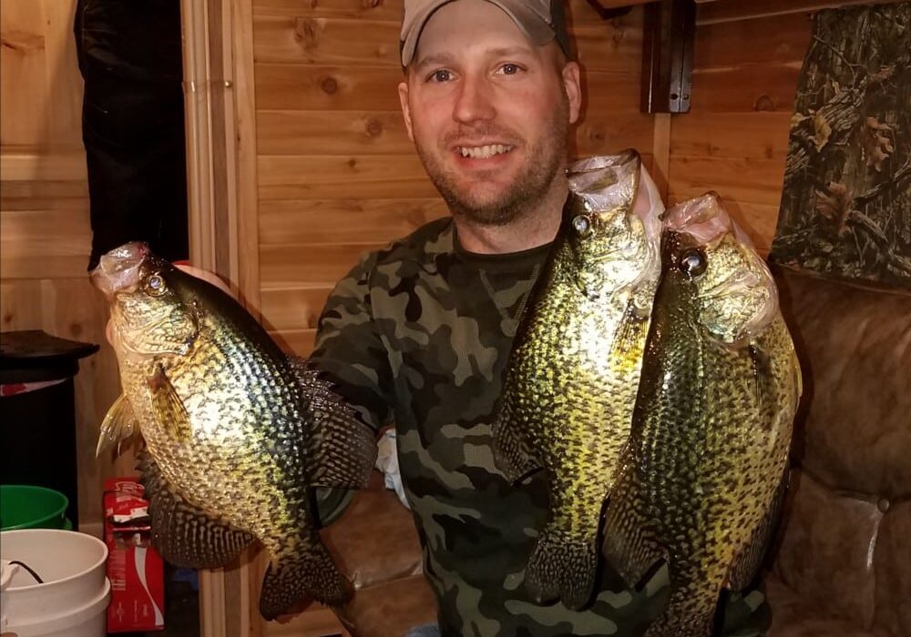 A smiling man in a camouflage shirt and baseball cap sits indoors, proudly holding up four large crappies—two in each hand. The room’s wooden walls create a cozy, cabin-like atmosphere.