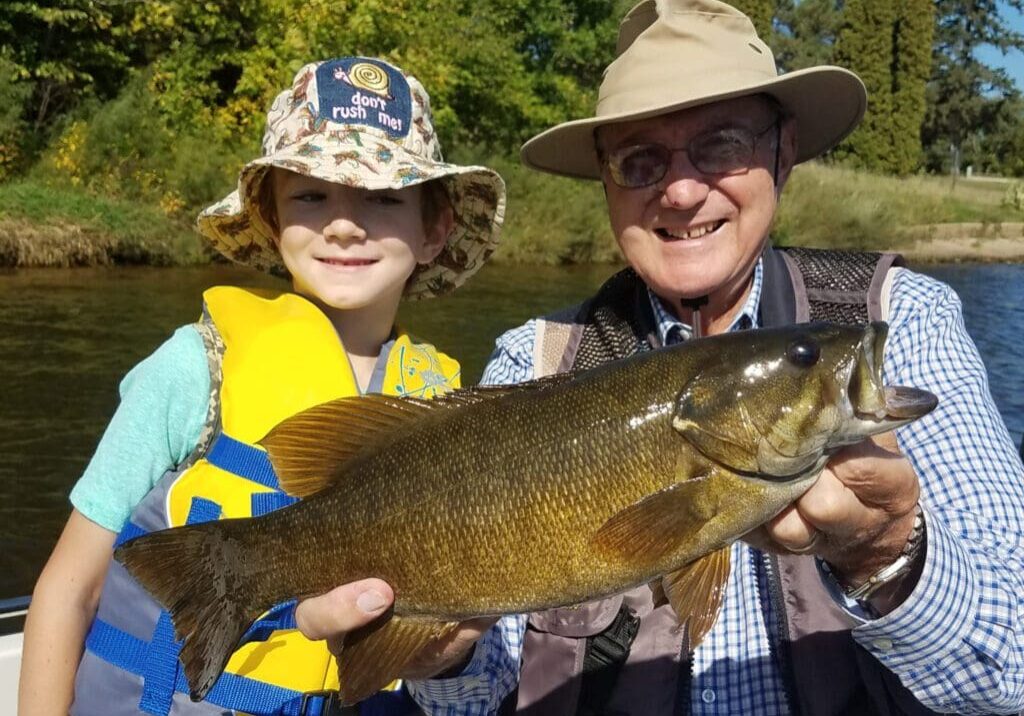 A smiling child in a yellow life jacket and an older man in a sun hat proudly hold a large fall fish together on a sunny day near a river, with green trees in the background.