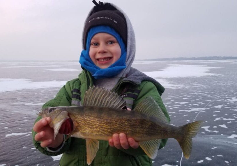 Smiling child in winter clothing holds a freshly caught fish while standing on a frozen, snow-dusted lake, proud of their December catch and ice report.