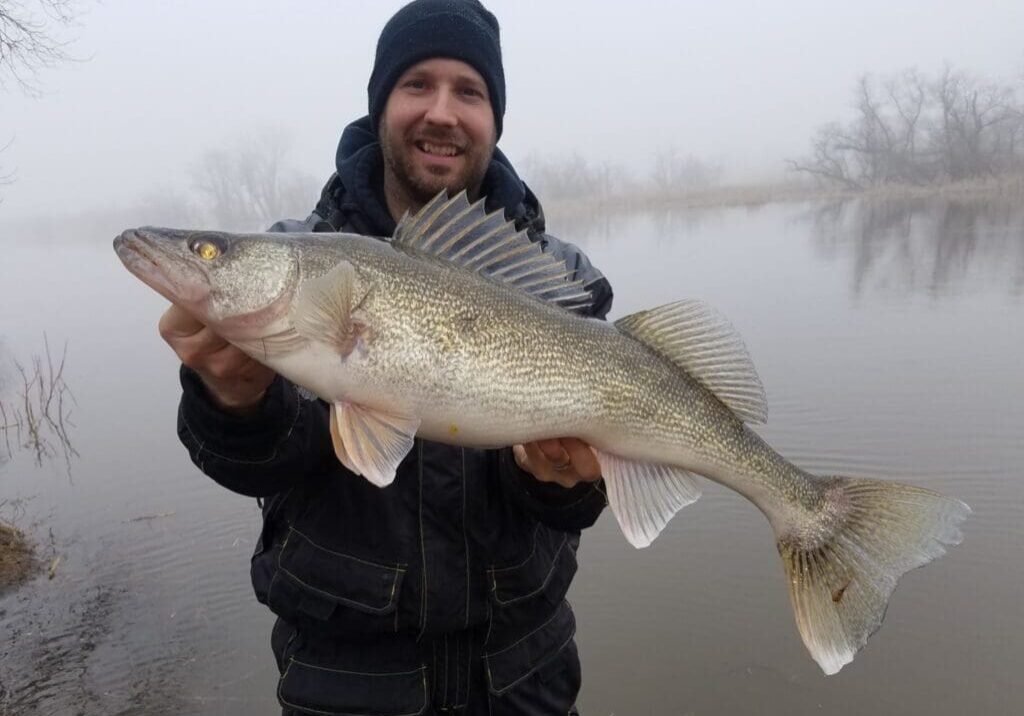 A person wearing a black beanie and jacket smiles while holding a large fish by the calm, foggy river in Alexandria, with bare trees in the background.