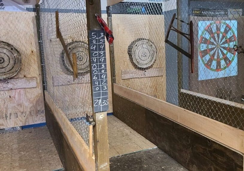 Woodsman Axe offers family friendly axe throwing lanes with wooden targets and chalk scoreboards. A digital dartboard is projected on the far wall, all safely enclosed by chain-link fencing and sturdy wooden barriers.