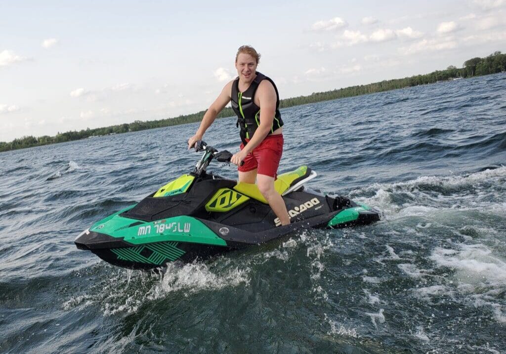 A person wearing a life jacket and red shorts is riding a green and black jet ski from Alexandria Jet Ski Rental on a lake, smiling at the camera with trees and a cloudy sky in the background.