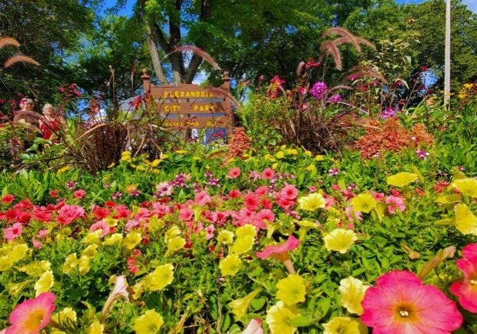 A vibrant flower bed with pink, yellow, and red blooms fills the foreground, with a wooden "Alexandria City Park" sign and lush green trees in the background under a partly cloudy blue sky.