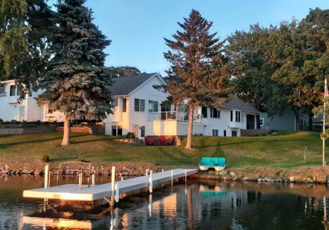 The Cottage on Lake Henry features a white house with large windows on a grassy hill by the lake, surrounded by trees. A small dock extends over the water as sunset light glows on the house, lawn, and docked rowboat.