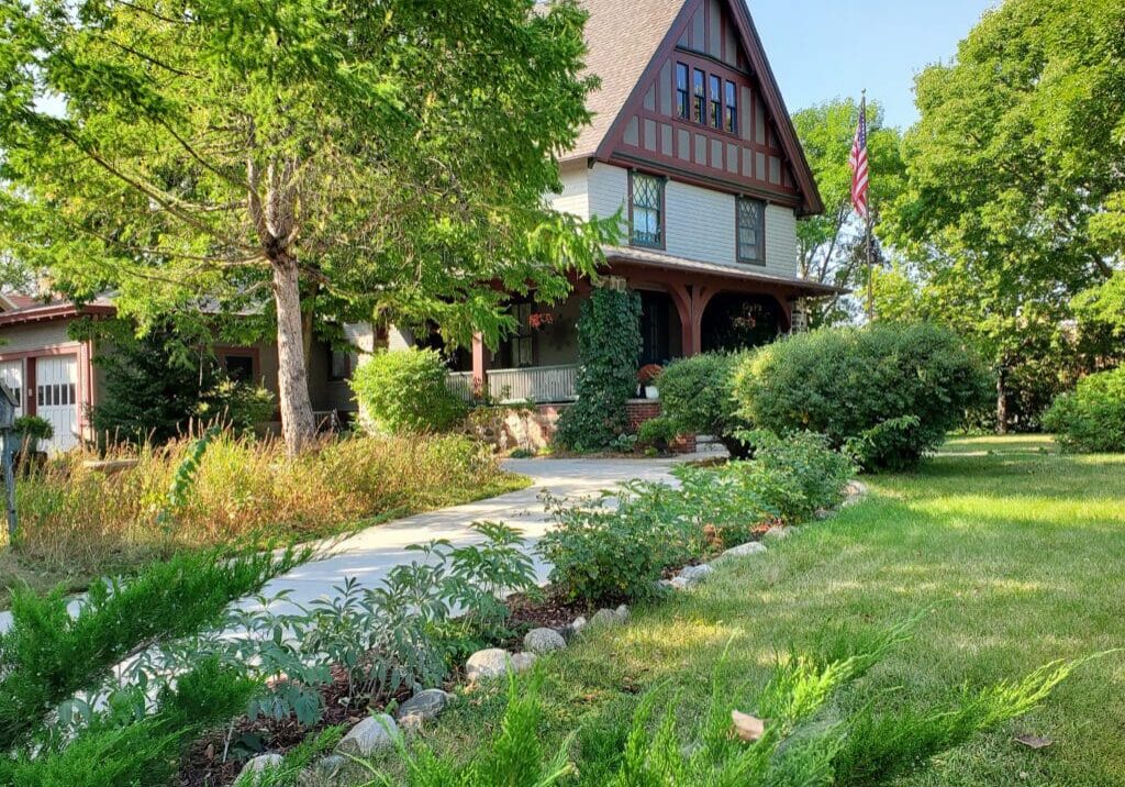 A large Tudor-style house, home to Cedar Rose Inn bed and breakfast Alexandria, sits behind a lush lawn and landscaped garden, surrounded by mature trees on a sunny day. An American flag is displayed near the front porch.